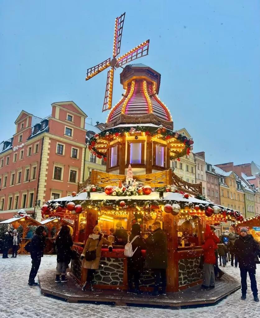 Windmill-shaped stall lit up and dusted with snow at Wrocław's Christmas market, in front of painted town houses